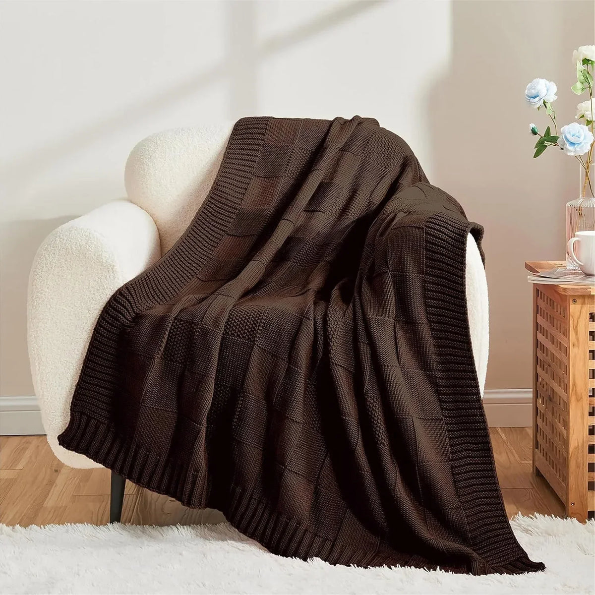 Couverture tricotée en laine de couleur marron foncé, posée sur un fauteuil blanc dans un intérieur cosy et accueillant.