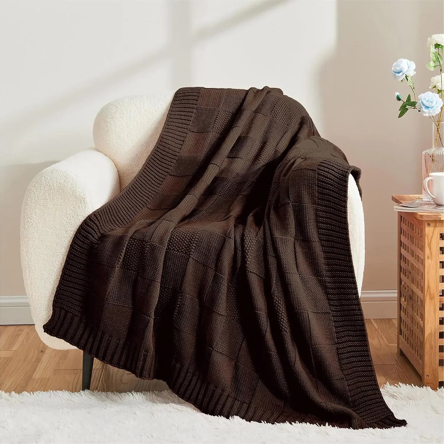 Couverture tricotée en laine de couleur marron foncé, posée sur un fauteuil blanc dans un intérieur cosy et accueillant.
