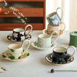 Une collection de tasses et soucoupes en céramique décorées de motifs floraux colorés, disposées sur une surface blanche avec une plante en arrière-plan.