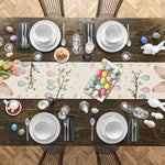 Table dressée pour Pâques avec une nappe ornée de motifs floraux et d'œufs colorés, entourée d'assiettes, de verres, de couverts et de décorations de Pâques.
