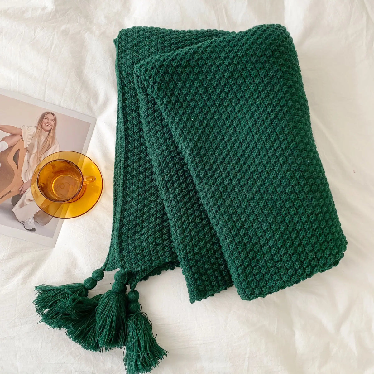 Une couverture tricotée verte émeraude avec des glands décoratifs sur un fond blanc avec une image de femme et une tasse en verre ambrée.
