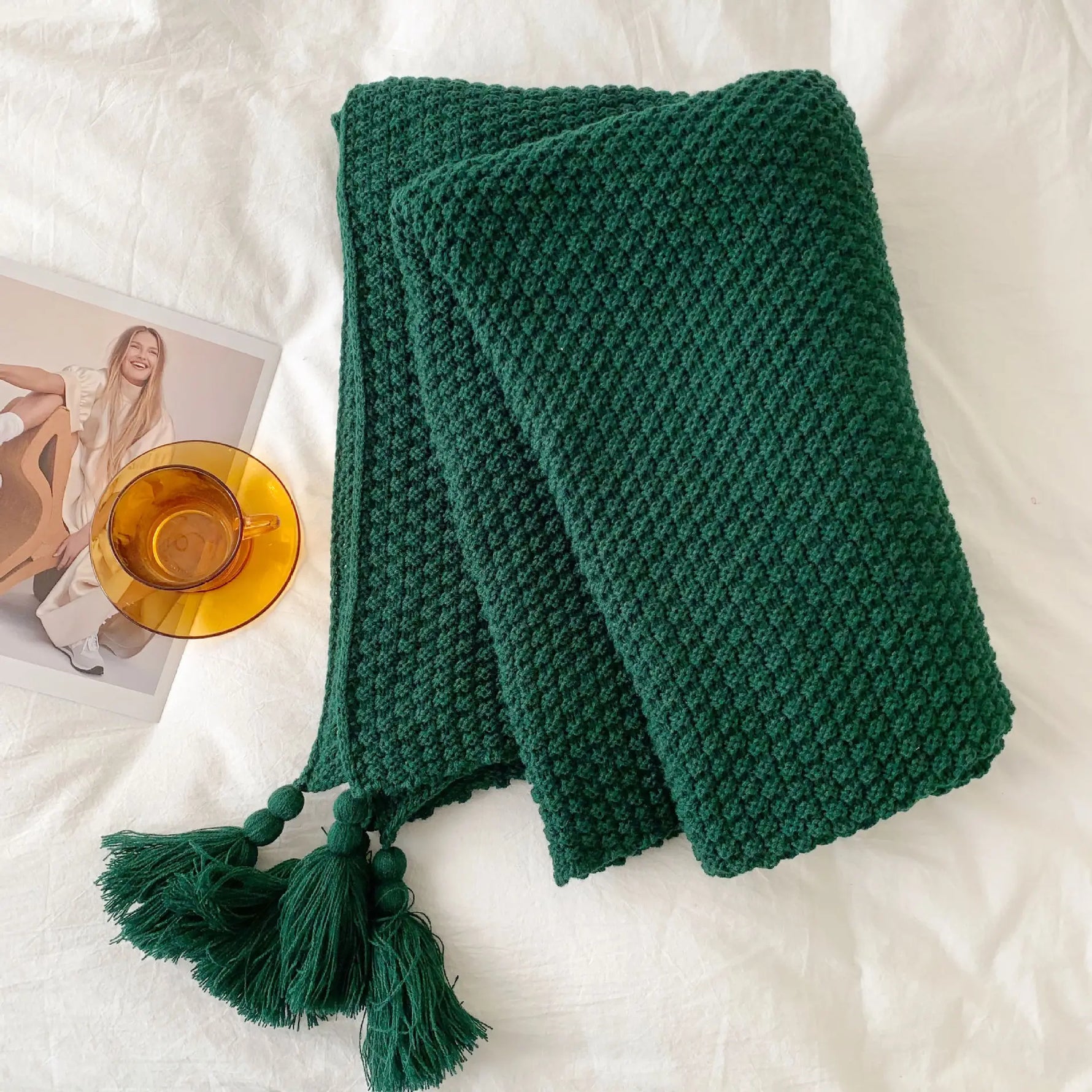 Une couverture tricotée verte émeraude avec des glands décoratifs sur un fond blanc avec une image de femme et une tasse en verre ambrée.