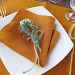 Une serviette de table en lin doré avec un bouquet de plantes vertes joliment attaché avec de la ficelle sur un plat blanc, créant une présentation élégante et naturelle.