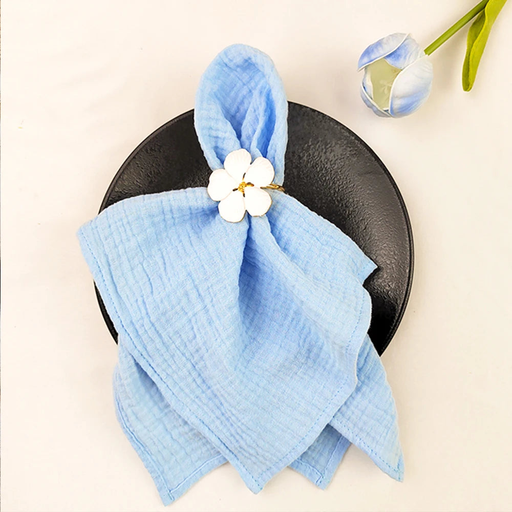 Une serviette en lin bleu ciel avec une fleur blanche épinglée dessus, posée sur une assiette noire, avec des tulipes en arrière-plan.