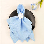 Une serviette en lin bleu ciel avec une fleur blanche épinglée dessus, posée sur une assiette noire, avec des tulipes en arrière-plan.