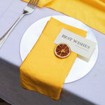 Une nappe et une serviette de table jaunes accompagnent une assiette blanche contenant un carton indiquant "BEST WISHES" ainsi qu'une tranche d'orange séchée comme décoration.