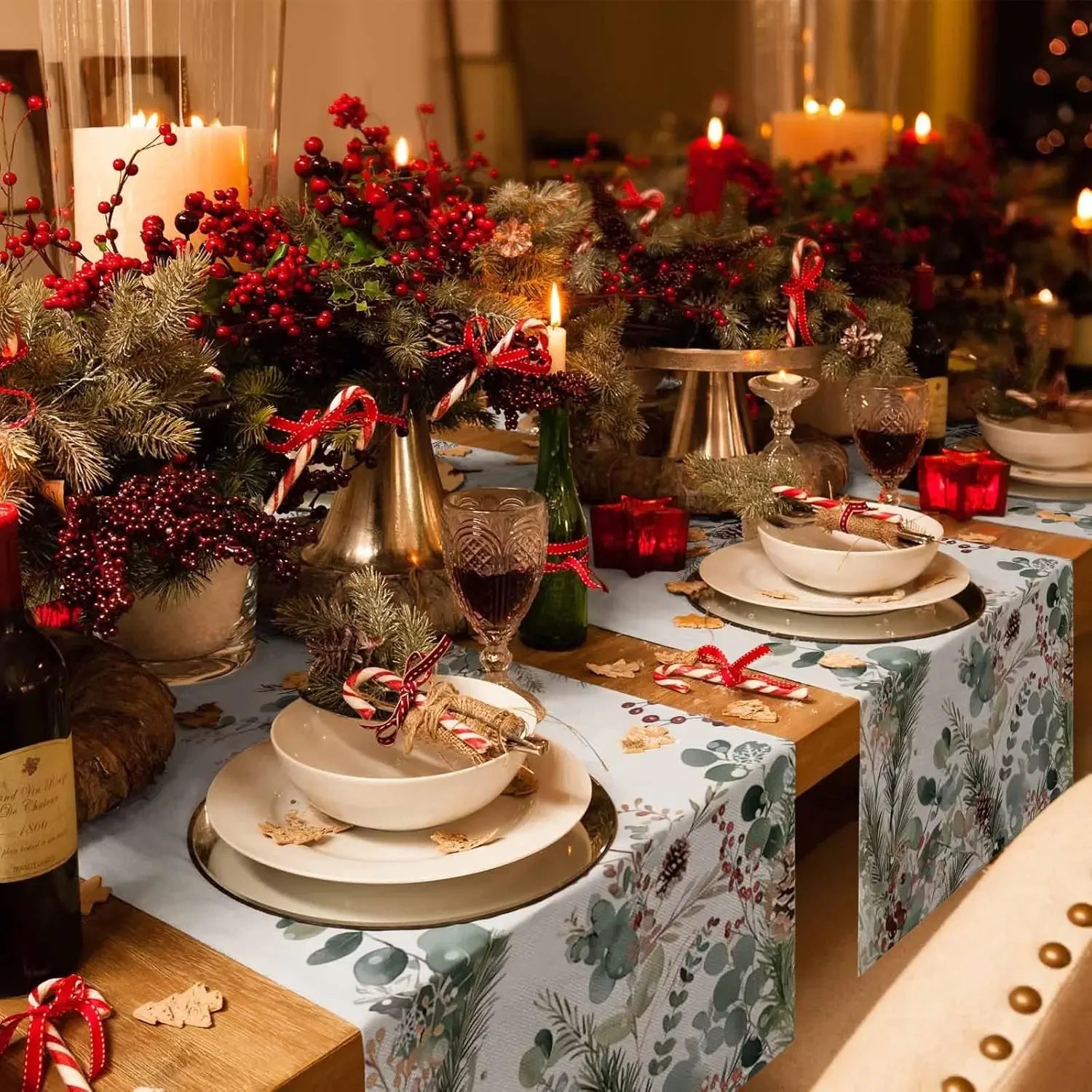 La table de fête est joliment décorée avec des branches de sapin, des baies rouges, des bougies et des verres à pied. Les assiettes en porcelaine blanche et le chemin de table fleuri créent une ambiance festive et chaleureuse.