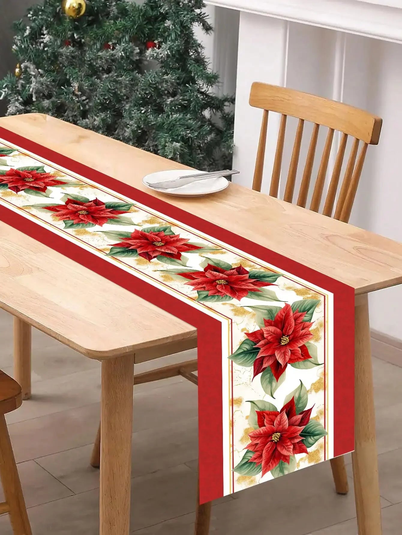 Cette image montre un chemin de table décoratif de Noël, avec des motifs de poinsettia rouges et verts sur un fond blanc, placé sur une table en bois avec une chaise en bois à côté.