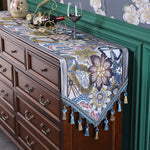 Ce mobilier vintage élégant présente un plateau orné de motifs floraux colorés dans des tons bleu, jaune et beige, rehaussé d'un élégant support en métal doré pour une bouteille de vin et deux verres, offrant une ambiance chaleureuse et raffinée.
