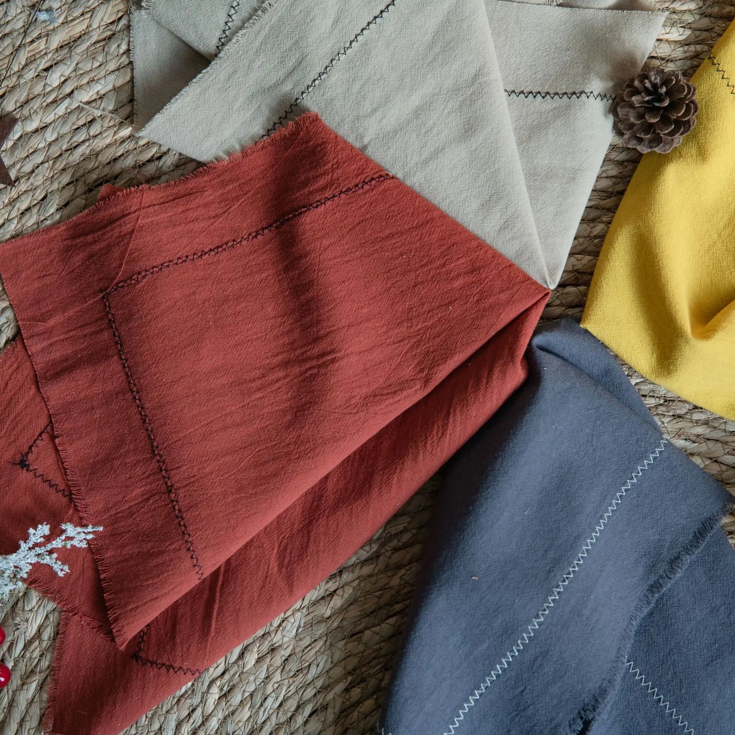 Une sélection d'articles textiles dans des tons chauds et automnaux, comprenant une couverture rouge, un plaid gris et une écharpe jaune, le tout posé sur un tapis tressé créant une ambiance cocooning.