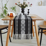 Une nappe noire et blanche avec des motifs ornementaux décore une table en bois avec des chaises noires, créant une ambiance bohème et élégante dans cette salle à manger.