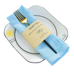 Sur une assiette blanche se trouve une serviette en tissu bleu ciel enroulée autour d'une fourchette dorée, avec un emballage jaune sur lequel est inscrit "Wanting 100% Cotton Cloth Napkin".