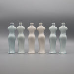 Six bouteilles en céramique blanche et bleu-gris alignées sur fond gris.