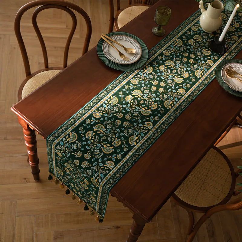 Nappe en coton élégante aux motifs floraux dorés sur fond vert foncé, ornée de pompons, posée sur une table en bois avec vaisselle et accessoires de décoration.