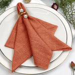 Serviette en lin de couleur orange disposée sur une assiette blanche, agrémentée de perles blanches sur un ruban doré.