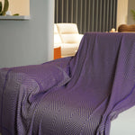 Couverture rectangulaire en coton de couleur violette et grise aux motifs géométriques décorée dans un salon avec des meubles beiges.