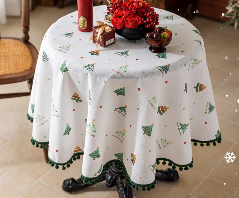Nappe de table ronde blanche imprimée avec des motifs de sapins de Noël, garnie d'une décoration florale rouge, de bougies et de petits objets décoratifs sur une table en bois dans un intérieur cosy.