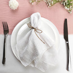 Une belle table dressée avec un linge de table blanc soigneusement plié et ligoté, entouré de fleurs blanches sur un fond rose.