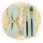 Couverts de table sur un set de table en paille tressée, avec une serviette de table et des couverts dorés sur un fond blanc.