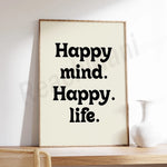 L'image montre un cadre en bois contenant le texte "Happy mind. Happy. life." en grands caractères noirs sur fond blanc. Le style du texte est simple et direct, transmetant un message de positivité et de bien-être.