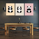 Affiches décoratives mignons avec des images de pandas assis sur un fond rose et beige, montées sur un mur gris foncé au-dessus d'une table en bois et de tabourets noirs, créant un bel ensemble décoratif pour une cuisine ou une salle à manger.