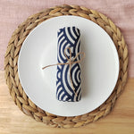 Serviette de table blanche et bleue rayée, attachée avec une ficelle, posée sur un napperon rond en fibres tressées.