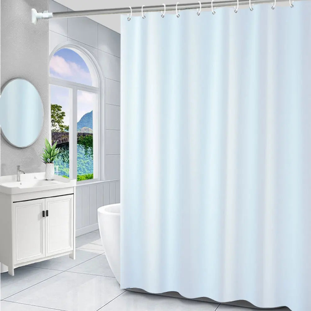 Salle de bain blanche et épurée, agrémentée d’un rideau de douche uni aux tons très clairs devant une large fenêtre offrant une vue sur un paysage.