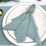 Des serviettes de table bleu-vert nouées avec de la ficelle naturelle, disposées sur une assiette blanche aux bordures dorées, entourées de branches de feuillage vert.