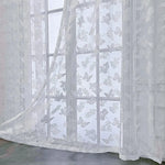 Voilage blanc transparent à motifs papillons et fleurs vu en gros plan dans une pièce lumineuse.