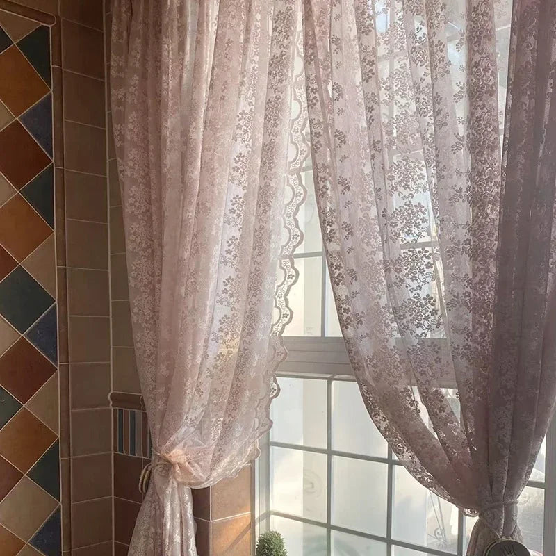 Voilage en dentelle rose clair à motifs floraux délicats, attaché partiellement pour laisser entrer la lumière naturelle dans une pièce carrelée chaleureuse.