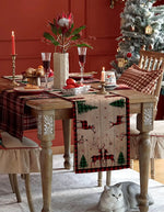 La table dressée pour un festin de Noël avec une nappe motif cerf, des bougies rouges, un bouquet de fleurs tropicales et une nappe à motifs forestiers crée une ambiance chaleureuse et festive, complétée par la présence d'un chat assis sur le sol.