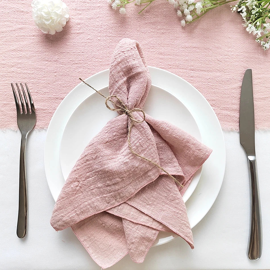 Cette image montre une jolie nappe de table rose pâle, avec une serviette pliée de manière originale et décorée d'un joli nœud sur une assiette blanche, accompagnée de couverts en métal sombre.