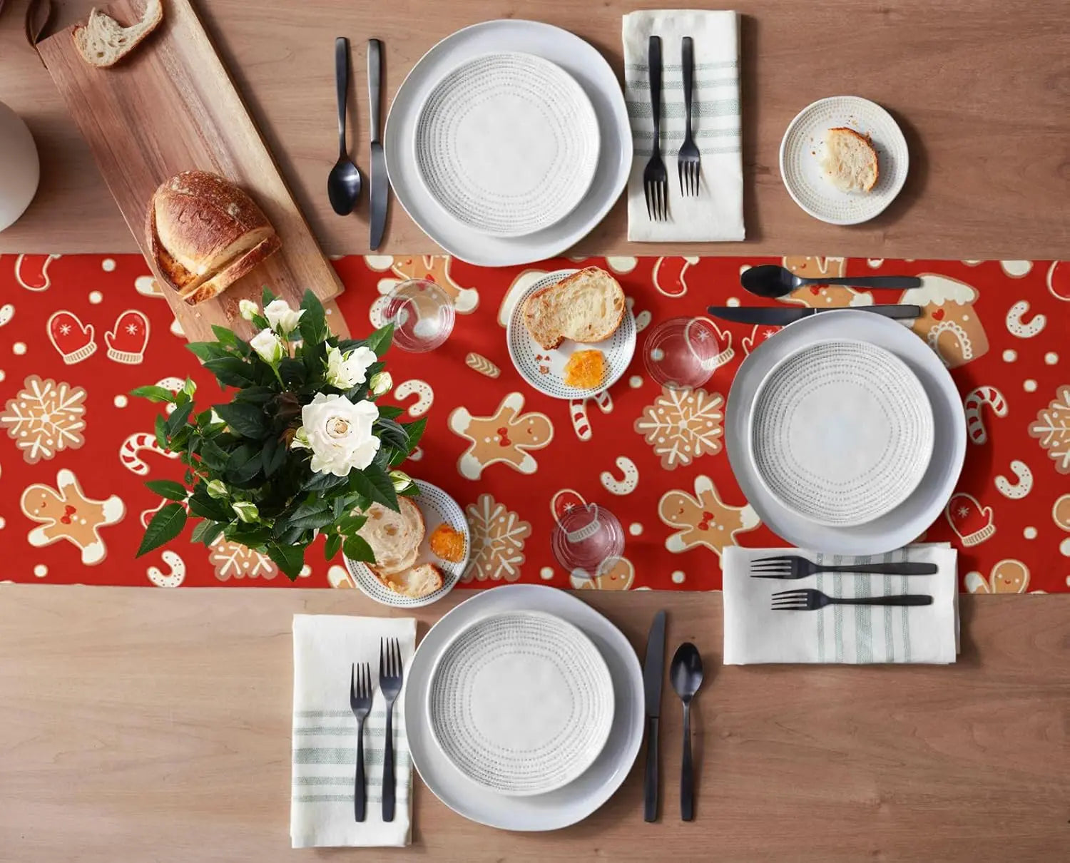 Une table de fête élégante avec de la vaisselle blanche, des ustensiles noirs et un napperon rouge festif décoré de motifs de bonhommes en pain d'épice et de flocons de neige.