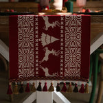 Ce tapis décoratif représente un motif géométrique abstrait en blanc sur un fond bordeaux, évoquant un style nordique ou scandinave, avec des franges et pompons en divers tons de marron, rouge et doré sur le pourtour.