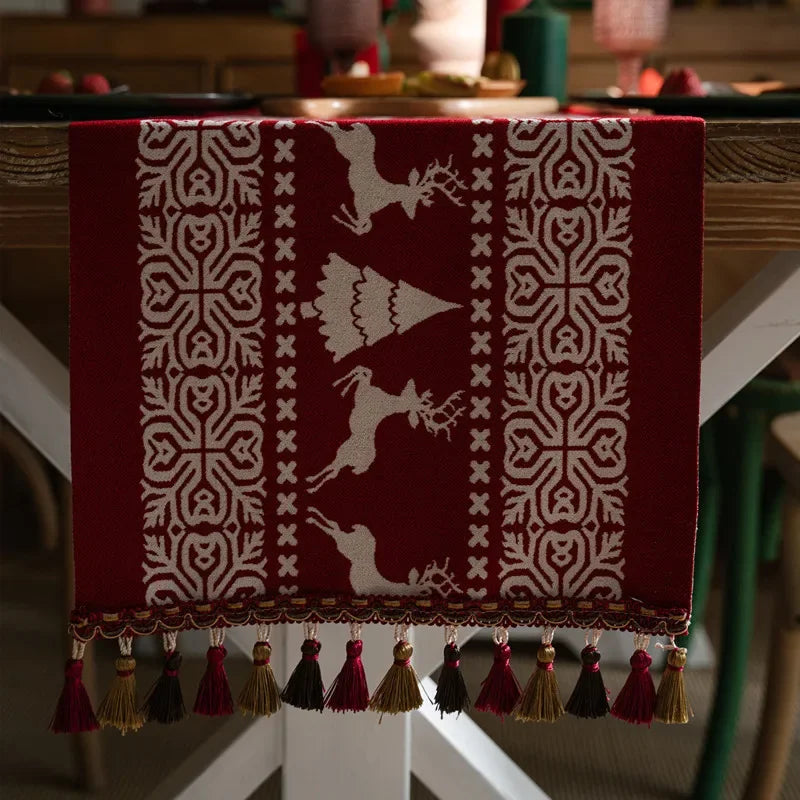 Ce tapis décoratif représente un motif géométrique abstrait en blanc sur un fond bordeaux, évoquant un style nordique ou scandinave, avec des franges et pompons en divers tons de marron, rouge et doré sur le pourtour.