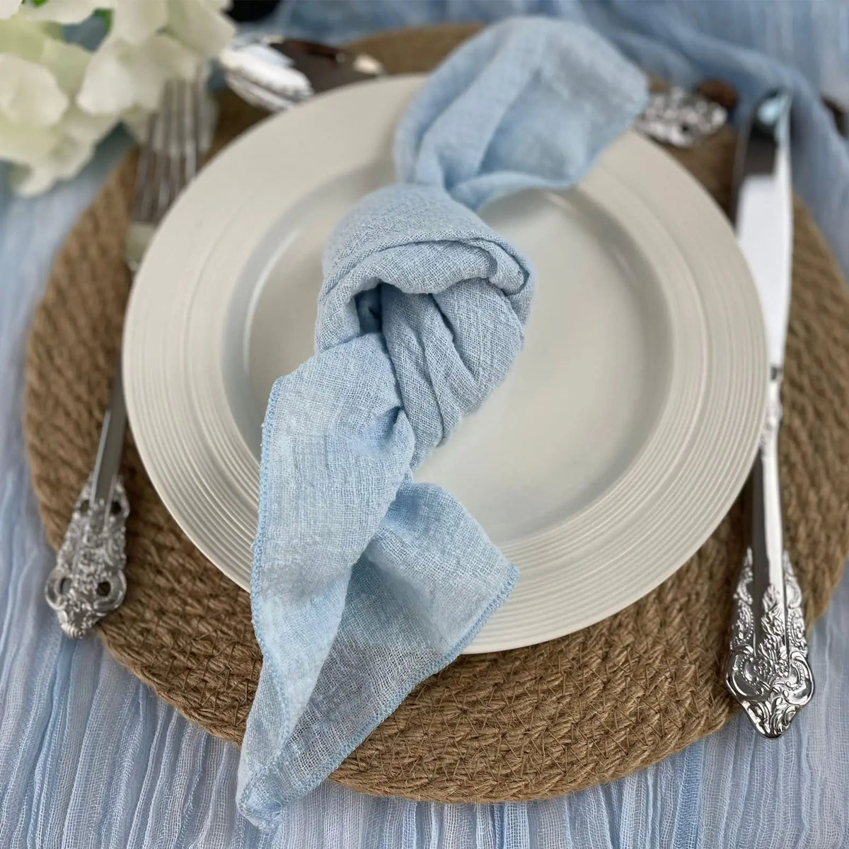 Serviettes en lin bleu clair posées sur des assiettes blanches et des couverts argentés sur un napperon en osier sur une table en bois.