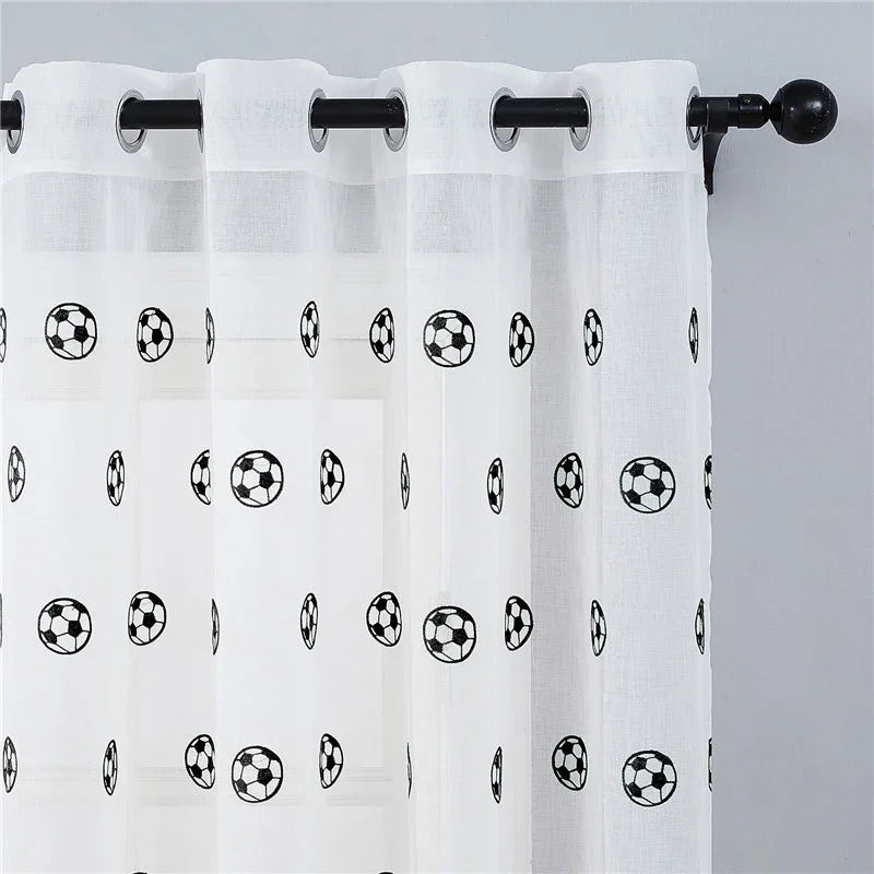 Voilage à motifs football suspendu sur une tringle noire, accroché à une fenêtre sur fond de mur gris.