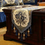 Un chemin de table en tissu brodé bleu et beige, avec des motifs floraux élégants et des franges, reposant sur un meuble en bois foncé.