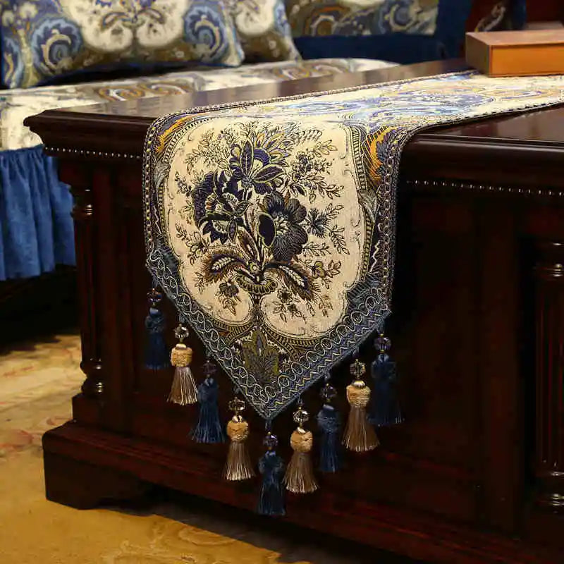 Un chemin de table en tissu brodé bleu et beige, avec des motifs floraux élégants et des franges, reposant sur un meuble en bois foncé.