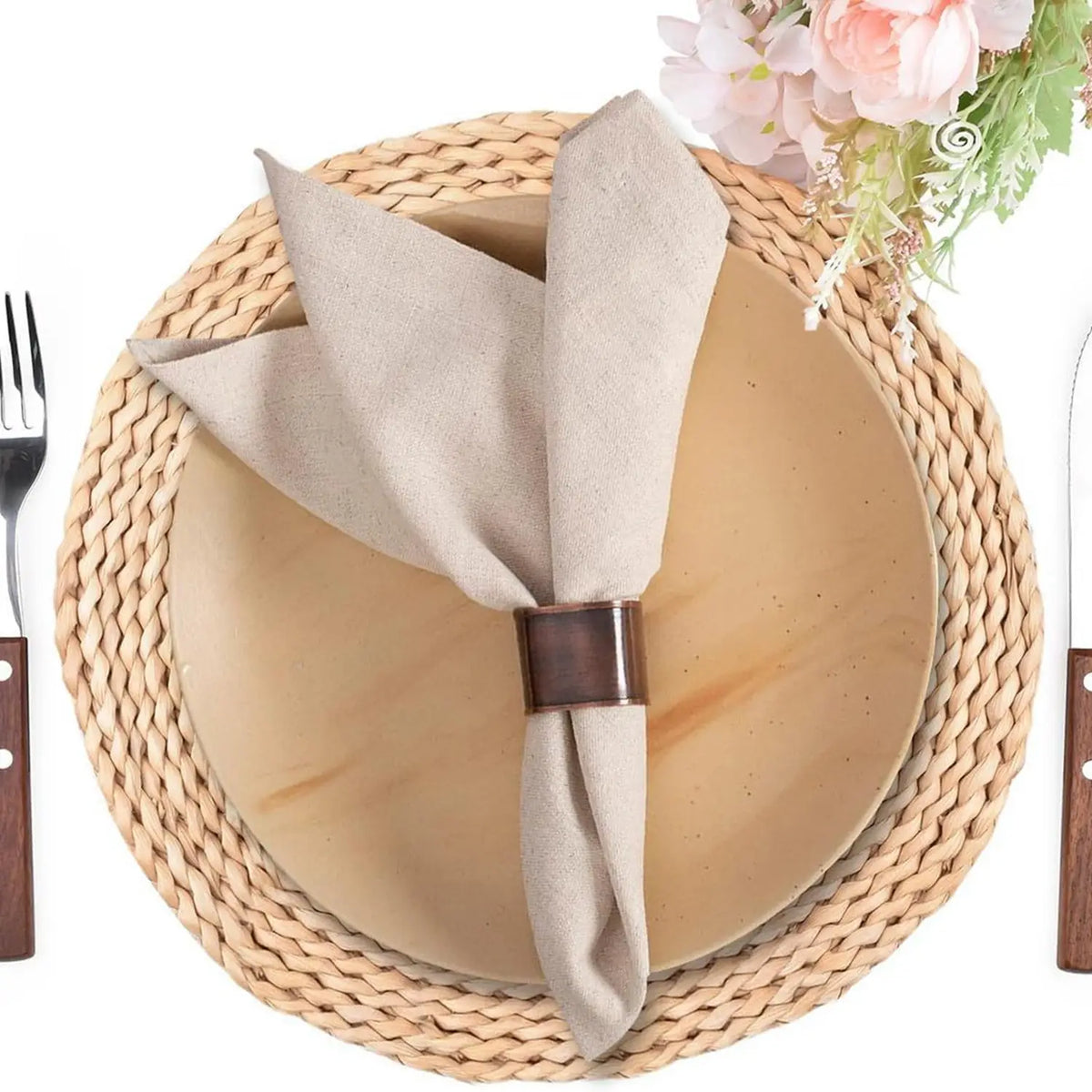 Un réglage de table élégant avec un napperon en osier, un linge beige et des couverts en métal, rehaussé d'un bouquet de fleurs roses.