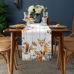 Une nappe de table en tissu avec des motifs de renards, d'écureuils et de citrouilles, entourée de verres à pied et d'un bouquet de fleurs, sur une table en bois dans un environnement douillet et automnal.
