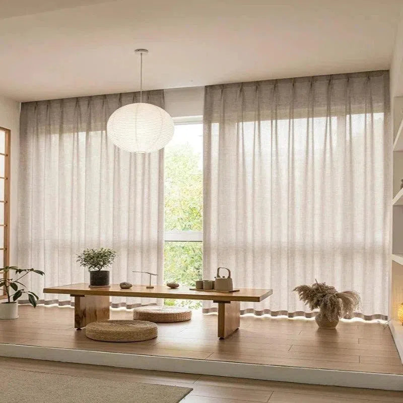 Salon minimaliste avec grandes fenêtres et rideaux modernes, table basse en bois élégante, décoration zen avec des plantes et coussins. Idéal pour créer un espace de détente lumineux et épuré.