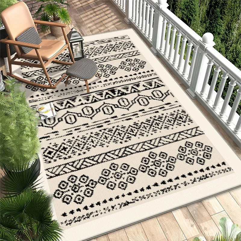 Tapis extérieur en fibres naturelles avec motifs géométriques en noir et blanc, posé sur un plancher en bois sur un porche avec des plantes tropicales.