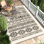 Tapis extérieur en fibres naturelles avec motifs géométriques en noir et blanc, posé sur un plancher en bois sur un porche avec des plantes tropicales.