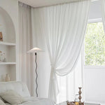 Voilage blanc noué délicatement au centre, dans un salon lumineux avec fauteuil blanc, lampe design noire et vue sur un jardin verdoyant.