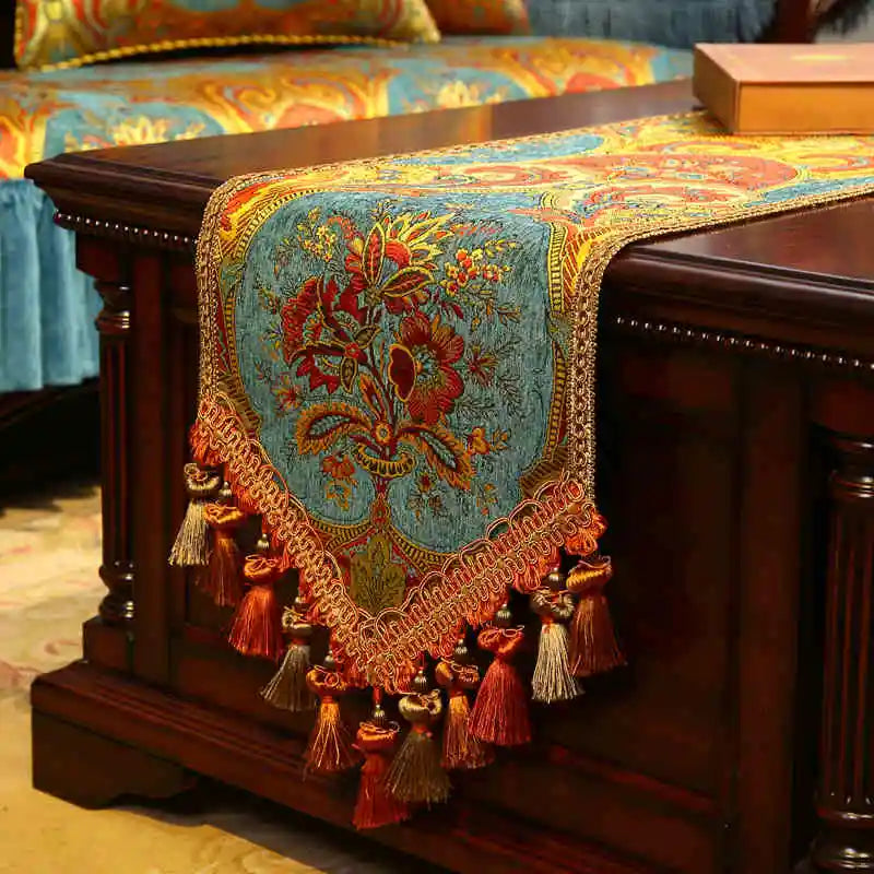 Un magnifique chemin de table en velours brodé de fleurs colorées aux couleurs chaudes et vibrantes, agrémenté de pompons et de glands rouges, créant une atmosphère chaleureuse et élégante.