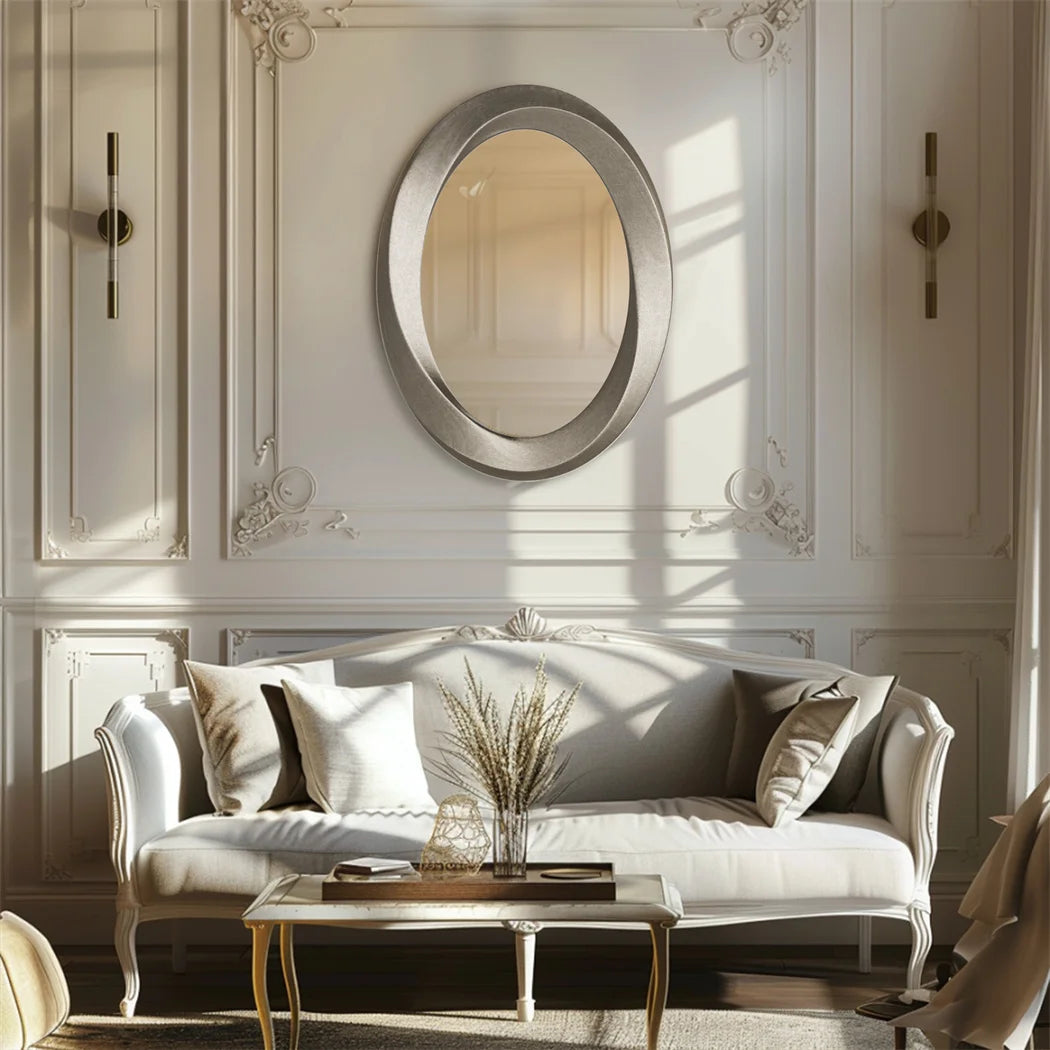 Élégant salon avec un miroir ovale argenté encadré sur un mur blanc orné de moulures, un canapé blanc à l'aspect raffiné, une table basse en métal doré et des éléments décoratifs harmonieux.