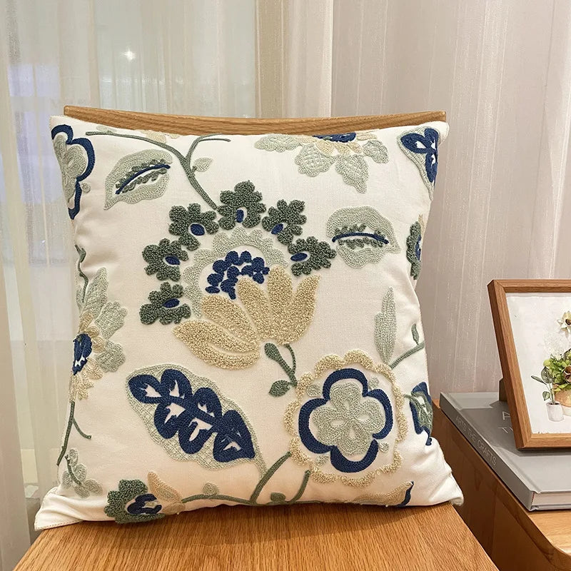 Coussin décoratif brodé avec des motifs floraux vintage bleu marine, crème et beige sur un fond blanc cassé.