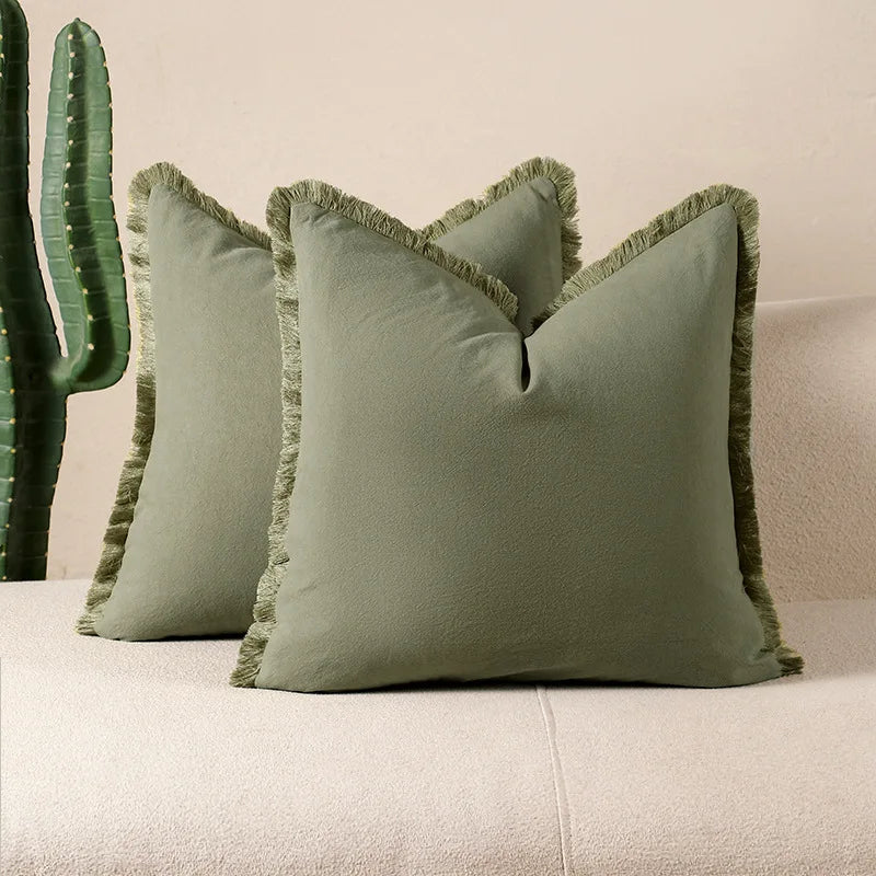Deux oreillers verts en velours doux avec des bordures effilées, posés sur un canapé blanc, à côté d'un grand cactus vert.