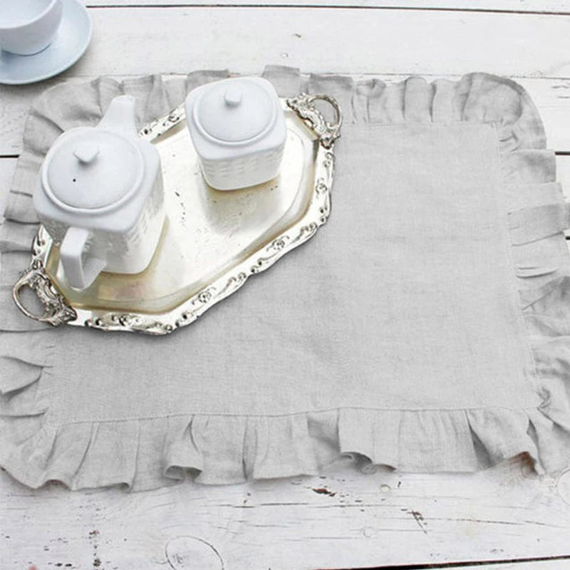 Sur une table en bois blanche, un service à thé vintage blanc et argenté avec une théière, une tasse et un plat décoratif, le tout sur un napperon en lin gris.